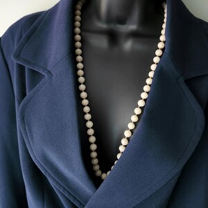 May include: A long strand of round, cream-colored beads worn over a dark navy blue blazer with a notched lapel. The necklace features a uniform series of smooth, spherical beads strung together, resting against the textured fabric of the jacket.