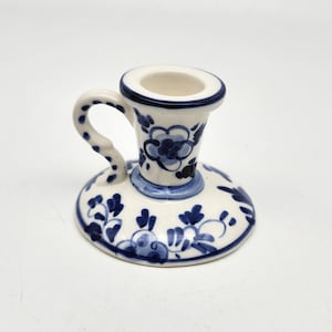May include: White ceramic candle holder with blue floral designs and a curved handle. The candle holder has a circular base and a cylindrical cup to hold a candle. The blue floral pattern decorates the base and the cup.
