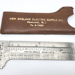 May include: A stainless steel Burndy Wire-Mike gauge with measurements and a brown leather holder. The holder has the text "NEW ENGLAND ELECTRIC SUPPLY CO." and other details. The gauge measures in inches.