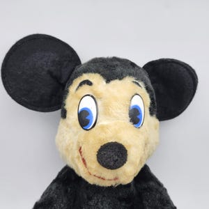 May include: A plush toy mouse with large black ears, a tan face, and a black nose. The mouse has large white eyes with blue irises and a red stitched smile. The toy is made of soft, plush material.
