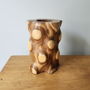 May include: A wooden vase with a unique, textured design. The vase is made of natural wood and has a brown and light brown color scheme. The vase is cylindrical in shape and has a smooth, rounded top.
