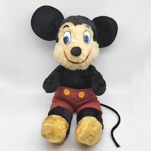 May include: Vintage Mickey Mouse plush toy. The plush features black fur, large black ears, a cream-colored face, red shorts with yellow buttons, and yellow feet. The toy has blue eyes and a smiling red mouth. A black tail extends from the back.