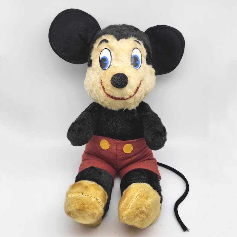 May include: Vintage Mickey Mouse plush toy. The plush features black fur, large black ears, a cream-colored face, red shorts with yellow buttons, and yellow feet. The toy has blue eyes and a smiling red mouth. A black tail extends from the back.