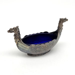 May include: A silver-toned Viking ship-shaped dish with a deep blue glass interior. The ship features dragon-head prows and decorative circular patterns along the sides. The dish is likely made of metal and glass.