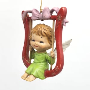May include: A vintage Christmas ornament featuring a cherubic angel in a green outfit, seated on a red swing shaped like a lyre. The ornament has a pink bow at the top and small white wings. The angel has blonde hair and a smiling face.
