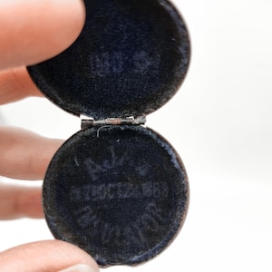May include: Open, round, antique jewelry box with a dark blue velvet interior. The box is made of a metal with a tarnished finish. The hinge is visible, and the box is held open by a hand.