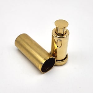May include: A gold-colored metal lipstick-style case with a removable cylindrical cap. The base features a push-button mechanism at the top with a scalloped edge, while the main body is smooth and cylindrical, showing a hollow interior.