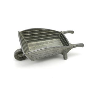 May include: A small, silver-colored wheelbarrow with a single wheel and two handles. The wheelbarrow has a rectangular bed with horizontal ridges. The object is made of metal and appears to be a miniature decorative item.