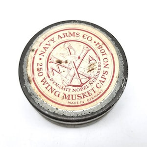 May include: A round, metal tin with a red and white label. The label features the text "NAVY ARMS CO." and "WING MUSKET CAPS." The tin has a distressed appearance, with visible wear and tear. The label also includes an anchor and cannon design.