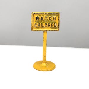 May include: A miniature yellow metal road sign featuring a rectangular top with the black embossed text WATCH CHILDREN. The sign is mounted on a thin vertical post attached to a circular base, all finished in a bright yellow painted coating.
