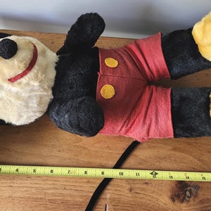 May include: A vintage Mickey Mouse plush toy, lying on a wooden surface. The plush features a black head and body, red shorts with yellow buttons, and yellow feet. A yellow measuring tape is visible.