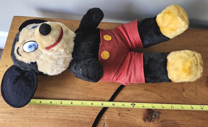 May include: A vintage Mickey Mouse plush toy, lying on a wooden surface. The plush features a black head and body, red shorts with yellow buttons, and yellow feet. A yellow measuring tape is visible.