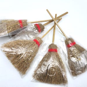 May include: Several miniature brooms with natural-colored bristles and wooden handles. Each broom is wrapped with a red band and packaged in a clear plastic bag. The brooms are arranged on a white surface.