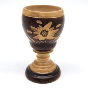 May include: A wooden goblet with a dark brown exterior and a light brown interior. The goblet features a carved floral design on the exterior. The base and stem are light brown, with a dark brown base.