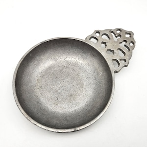 May include: A small, round, metal bowl with a decorative handle. The bowl is a dull gray color with a textured surface. The handle has a cut-out design. The bowl is likely used for serving or display.