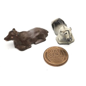 May include: Two vintage toy farm animals, a brown cow and a white and black cow, lying down. A copper penny is in the background.
