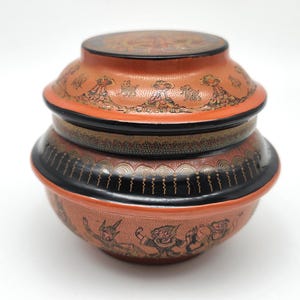 May include: A stacked, orange-red decorative container with a lid, featuring black and gold accents. The container is adorned with detailed illustrations of figures and patterns. The base has a rounded shape, and the lid is flat, creating a unique, artistic design.