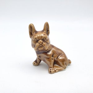 May include: A brown ceramic figurine of a French Bulldog in a seated position. The dog has a wrinkled face, upright ears, and a short tail. The figurine is a solid color and sits on a white surface.