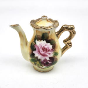 May include: A small, decorative porcelain teapot with a hand-painted rose design. The teapot features a yellow and gold color scheme, with a pink and white rose on the front. The handle and spout are gold-colored.