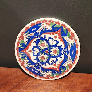 May include: A round, ceramic coaster with a detailed floral design. The coaster features a white base with a blue, red, and green pattern. The design includes stylized flowers and leaves, with a red border.