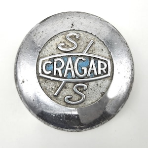 May include: A round, silver-toned metal emblem with the text "CRAGAR" in a white, stylized font, set against a blue background. The emblem also features the letters "S" and "TS" in a decorative script.