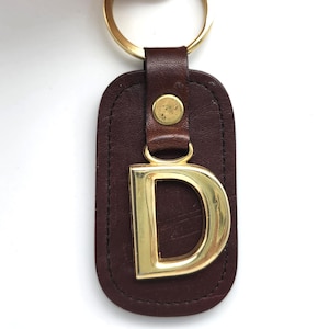 May include: A brown leather keychain with a gold-colored metal letter "D" and a gold-colored metal ring. The keychain has a stitched border and a small gold-colored button. The background is white.