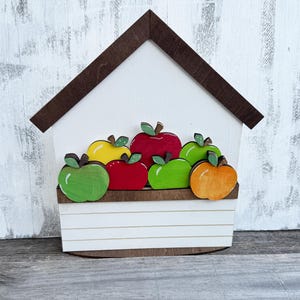 Può includere: Decorazione in legno a forma di casa, con tetto marrone e corpo bianco. La casa presenta una mensola con diverse mele di legno colorate nei toni del rosso, verde, giallo e arancione. Lo sfondo è una superficie bianca strutturata.
