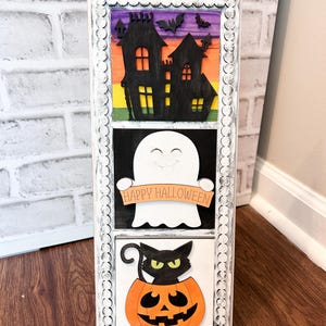 Puede incluir: Un marco de madera blanco con tres secciones separadas. La sección superior presenta una silueta negra de una casa con murciélagos volando por encima. La sección del medio presenta un fantasma blanco sosteniendo una pancarta que dice "Happy Halloween". La sección inferior presenta un gato negro sentado dentro de una calabaza naranja tallada.