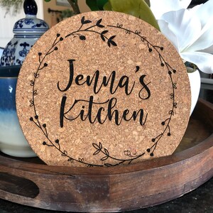 May include: A round cork trivet with a black floral wreath design and the text "Jenna's Kitchen" in a black cursive font.