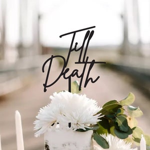 May include: A three-tiered wedding cake with black and white frosting. The cake is decorated with white flowers and greenery. A black topper with the words "Till Death" is on top of the cake.