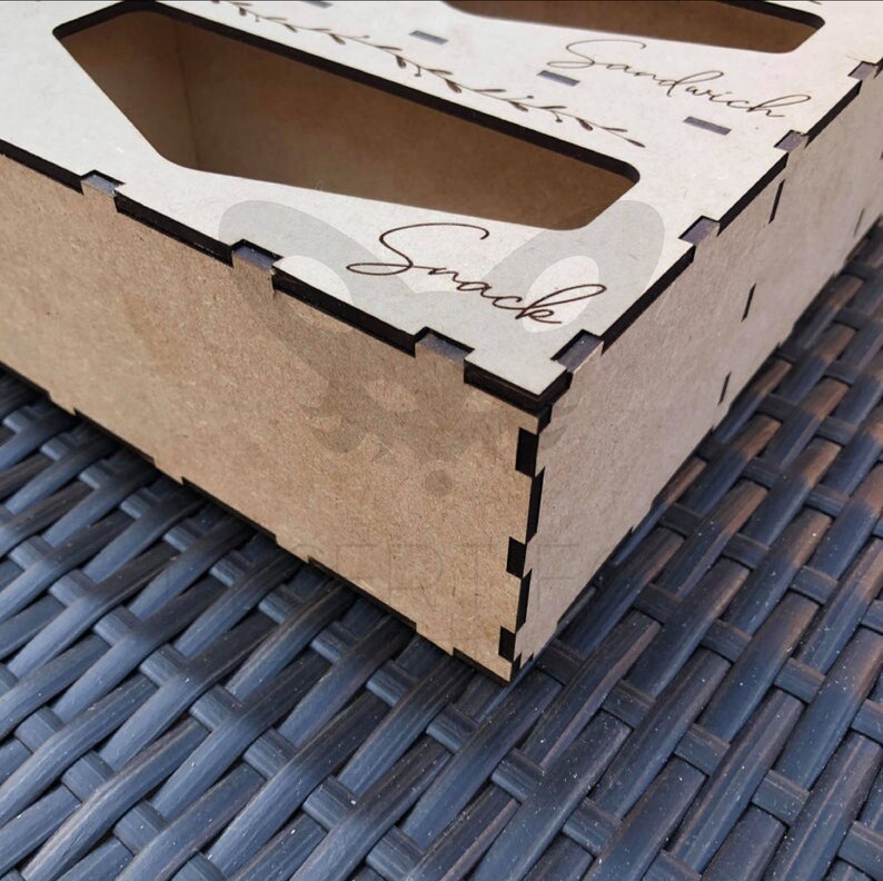 May include: A wooden snack and sandwich box with laser-cut compartments. The box is made of light brown wood with dark brown edges. The word "Snack" is written in cursive on the side, and "Sandwich" is written on the top.