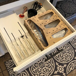 May include: A white kitchen drawer is open, revealing organized storage solutions. It contains a wooden organizer labeled "Gallon", "Quart", "Sandwich", and "Snack" for bags, along with measuring cups, spoons, and knives. The drawer is accented with black hardware.