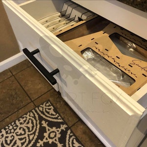 May include: Open white drawer with black handles. Inside, a knife block and a cardboard organizer with the word "Sandwich" are visible. The drawer is set against a tiled floor and a patterned rug.