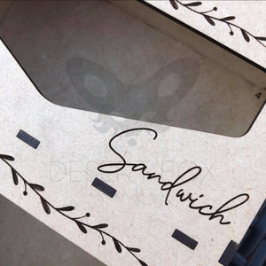 May include: A laser-cut wooden sandwich box with a clear window. The box is light brown with the word "Sandwich" in a cursive font. Decorative leaf and dot patterns are on the sides.