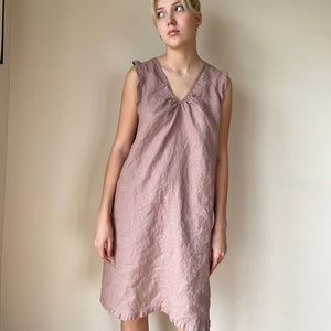 May include: A dusty pink linen dress with a v-neck and ruffled straps. The dress has a gathered detail at the neckline and a slightly A-line silhouette.
