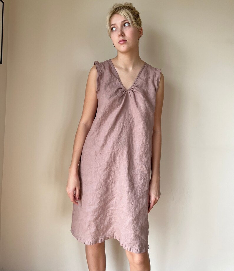 May include: A dusty pink linen dress with a v-neck and ruffled straps. The dress has a gathered detail at the neckline and a slightly A-line silhouette.