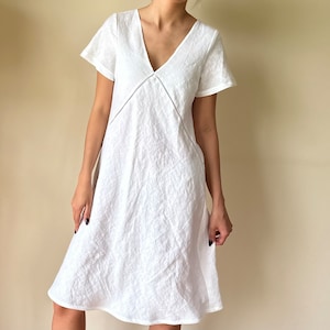 May include: A white linen dress with a V-neckline and short sleeves. The dress has a simple, relaxed fit and a slightly A-line silhouette.