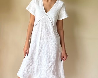 Puede incluir: Un vestido blanco de lino con escote en V y mangas cortas. El vestido tiene un corte sencillo y relajado y una silueta ligeramente acampanada.