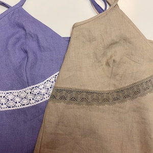 May include: Two sleeveless tops with lace trim. The top on the left is a light purple color with white lace trim. The top on the right is a light brown color with brown lace trim.