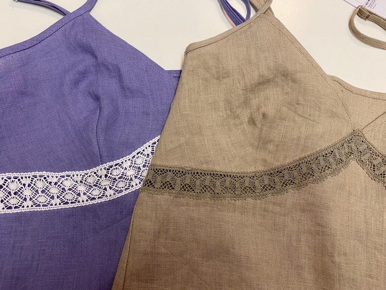 May include: Two sleeveless tops with lace trim. The top on the left is a light purple color with white lace trim. The top on the right is a light brown color with brown lace trim.