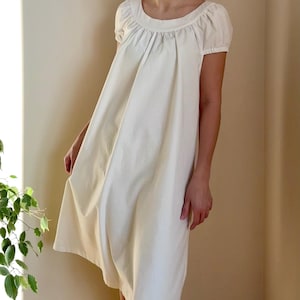 Pode incluir: Vestido creme com decote redondo e mangas curtas bufantes. O vestido tem uma silhueta solta e fluida e vai até a metade da coxa. O tecido parece ser um algodão ou linho leve, adequado para climas quentes.