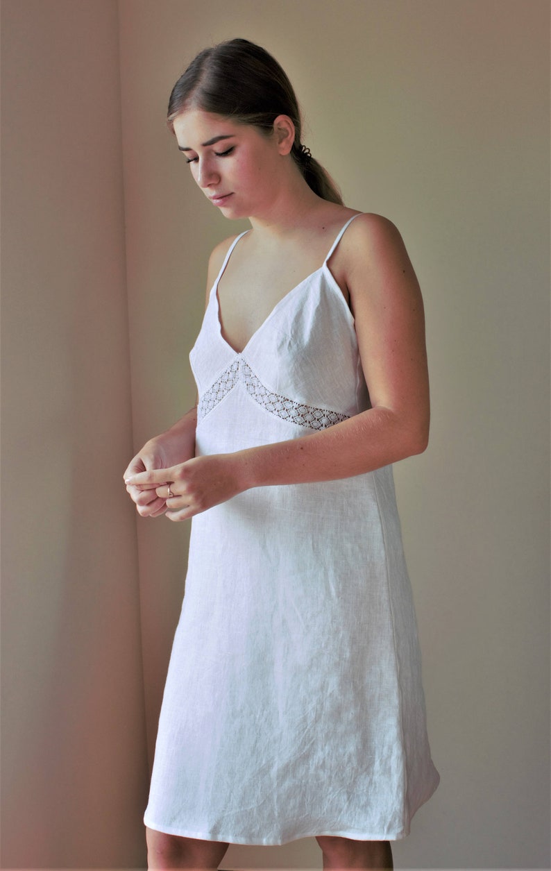 May include: A white linen slip dress with a delicate lace trim around the neckline. The dress has a simple, elegant design and a relaxed fit.