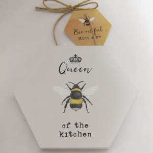 May include: White hexagonal sign with a black and yellow bee illustration and the text "Queen of the kitchen". A small tag with a bee illustration and the text "Bee-utiful Mini Sign" is attached to the sign.