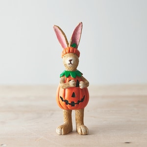 May include: A brown rabbit figurine dressed as a jack-o'-lantern for Halloween. The rabbit is holding a pumpkin with a carved face and wearing a green leaf collar.
