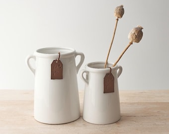 May include: Two white ceramic vases with small handles, one holding dried stems. Each vase has a brown tag that says "for you". The vases are on a light brown wooden surface.