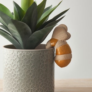 May include: A ceramic bee planter with a yellow and brown striped body, peeking out from a grey planter with a floral pattern. The planter is filled with a green succulent plant.