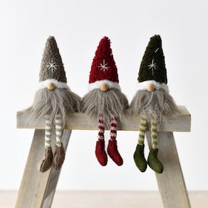 Puede incluir: Tres gnomos de peluche con largas barbas grises, sentados en un banco de madera. Cada gnomo tiene un sombrero de diferente color: gris, rojo y verde, cada uno con una estrella blanca. Sus piernas a rayas cuelgan, con zapatos del mismo color.