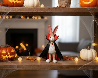 May include: A standing rabbit figurine dressed as a vampire, featuring long ears, a black cape with a high red collar, and a small red pendant around its neck. The rabbit has a light brown textured body and is positioned in a central upright stance.