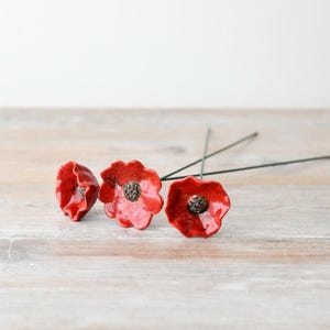 Puede incluir: Tres flores de amapola de cerámica roja con centros negros sobre tallos verdes delgados. Las flores están dispuestas en una fila sobre una superficie de madera clara.