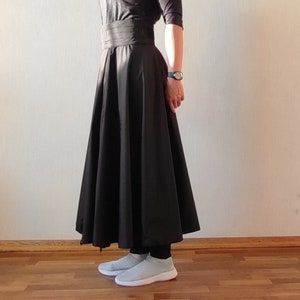 May include: A black, full-length skirt with a wide waistband. The skirt is paired with a black top and light gray slip-on sneakers. A watch is visible on the wrist. The skirt has a flared silhouette.
